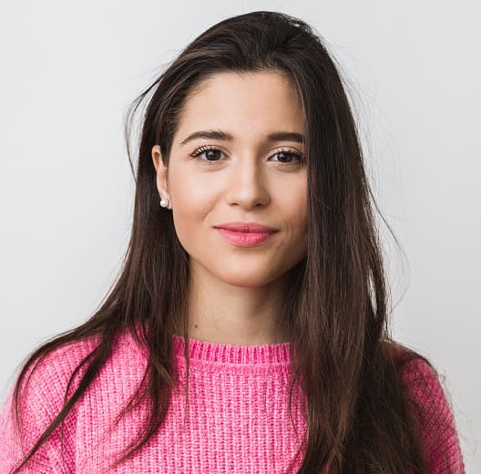 a smiling girl with long brown hair, brown eyes, pearl earrings in a bright pink sweater