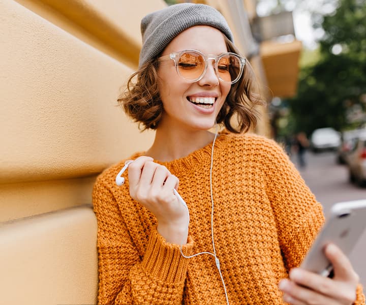Smiling young woman in an orange sweater and beanie holding a smartphone outdoors